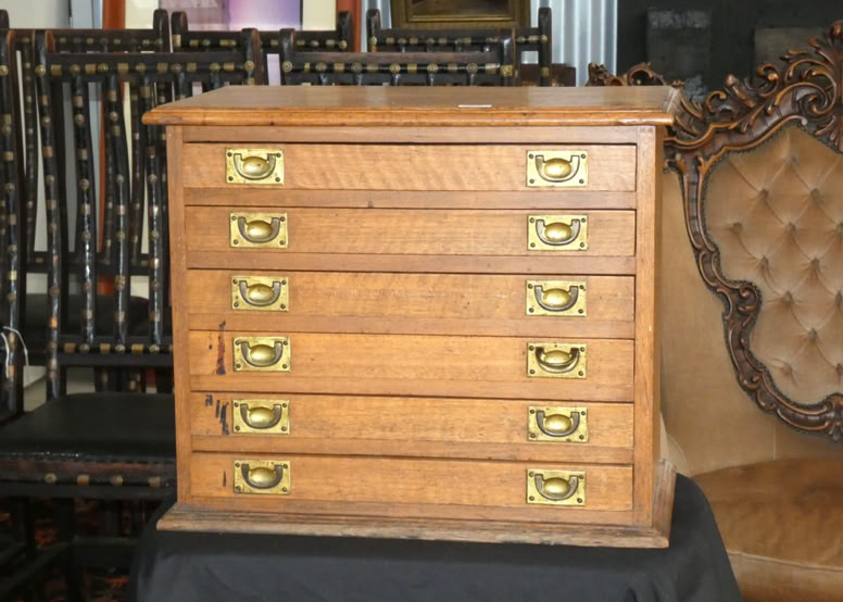 Wooden chest with six drawers and brass handles, placed on a dark cloth, with ornate chairs in the background.