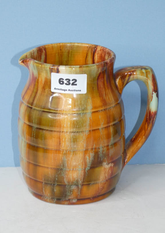 A ceramic pitcher with a multicolored glaze, featuring a handle and a spout, placed against a blue background. A label with the number 632 is attached to the pitcher.