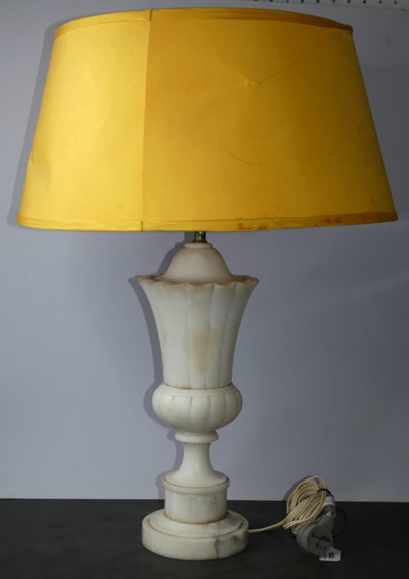 A table lamp with a yellow lampshade and a white, ornate base, placed on a surface with a coiled cord beside it.
