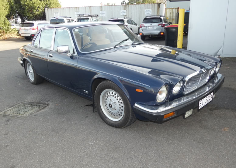 A dark blue classic sedan parked on a paved surface with other cars and a building in the background.