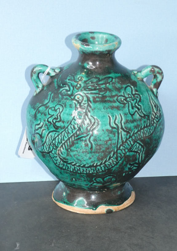 A green ceramic vase with intricate designs, featuring two small handles near the top, placed on a dark surface against a light blue background.